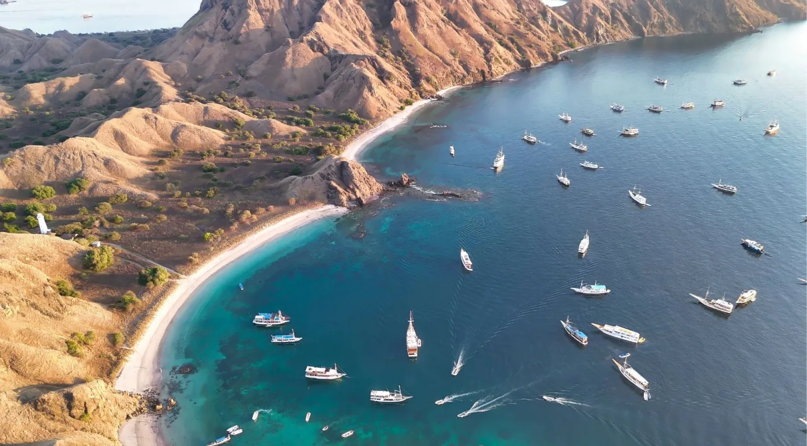 Aerial view of a curved beach on a dry hillside with yachts and small boats anchored in clear shallow water
