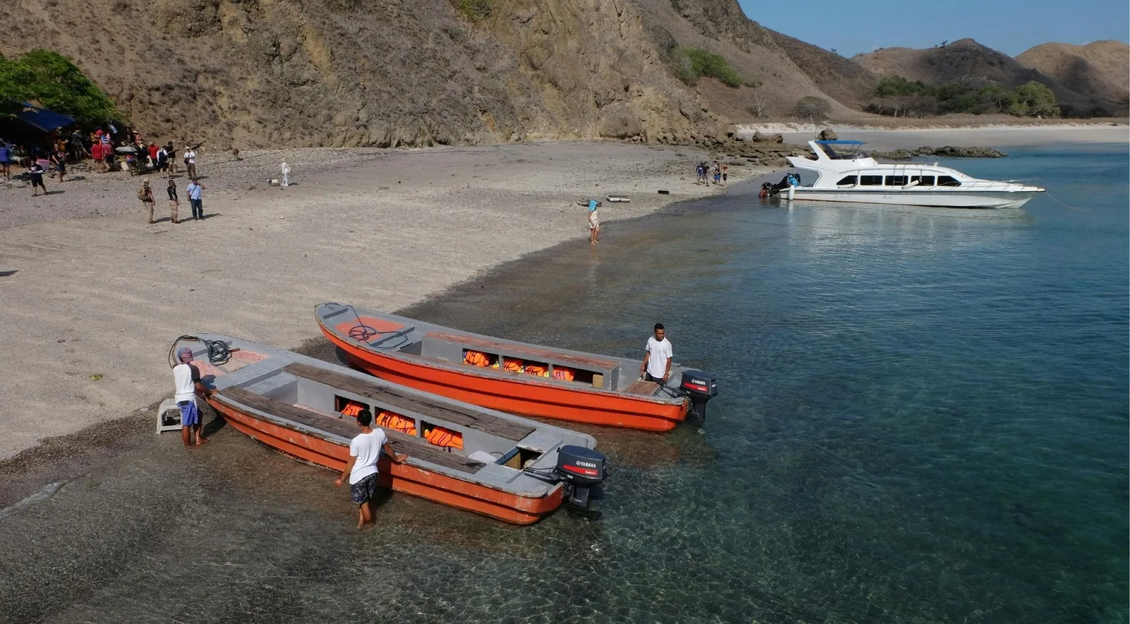 Speedboats and a covered transfer boat drawn up on a grey beach below the bare hills of Komodo National Park