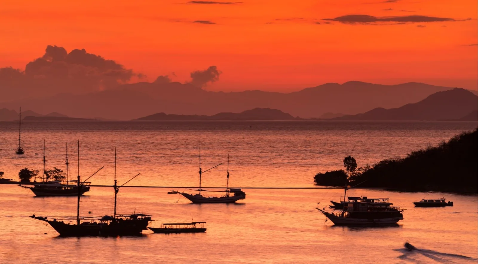Silhouettes of Phinisi sailing boats on still water beneath a deep orange Komodo sunset