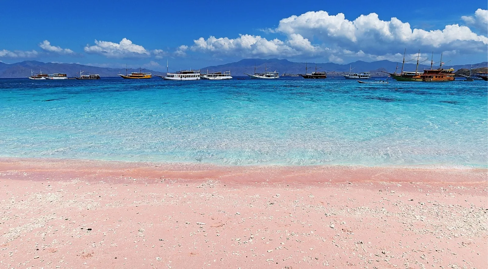 Pink sand at the waterline of Pantai Merah with day boats moored on the turquoise water beyond