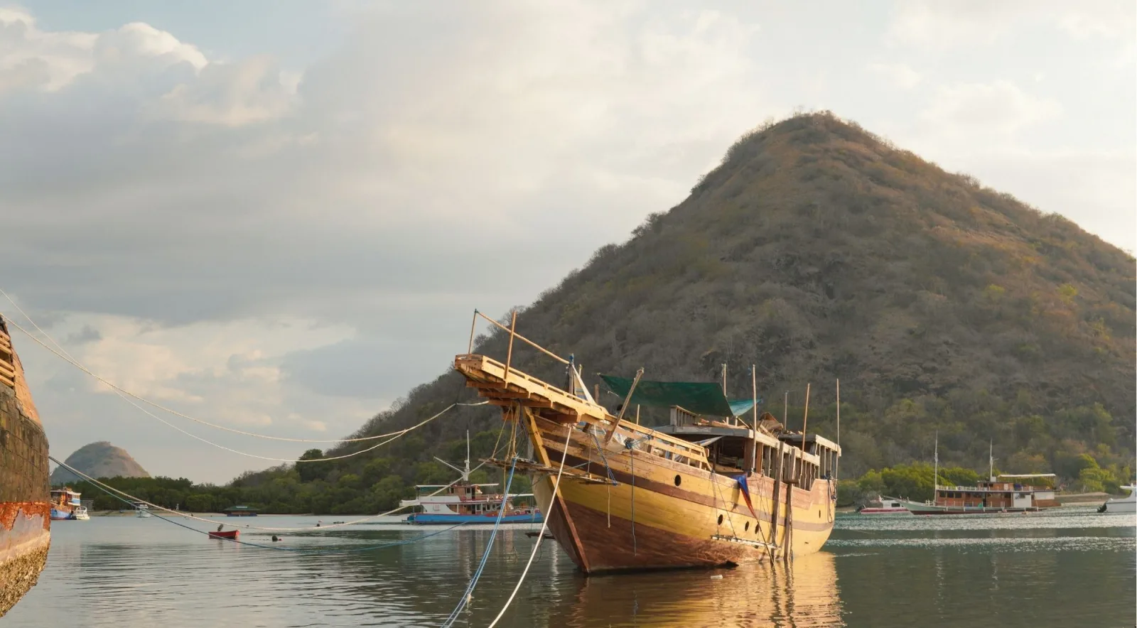A traditional wooden Phinisi at anchor in a still bay below a steep green headland